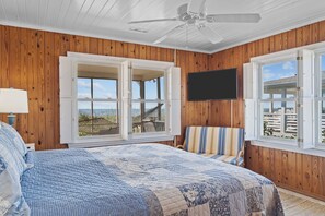 A bedroom with a bed, a chair, a TV, and large windows with a view of the beach. - Holliday Haven #200 - Oceanfront Escape in Topsail Beach, Sleeps 9! (Topsail Beach)