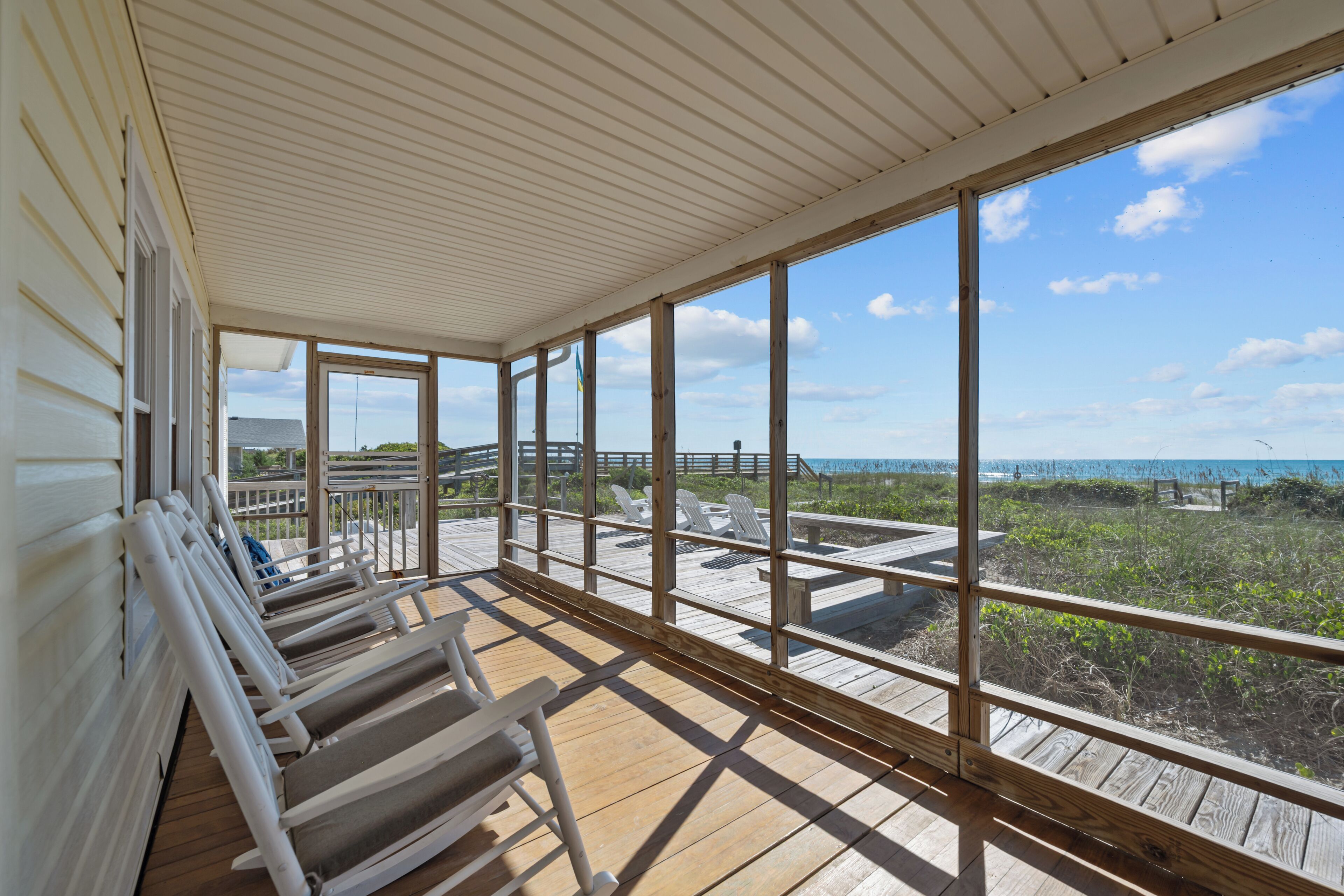 A covered outdoor deck with wooden railings and lounge chairs, overlooking a beach and ocean.