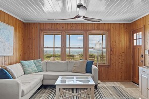 A room with a wooden interior, a beige sofa with patterned cushions, a glass-top coffee table, and a view of the beach through large windows. - Holliday Haven #200 - Oceanfront Escape in Topsail Beach, Sleeps 9! (Topsail Beach)