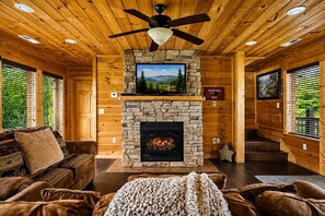 A log cabin interior with a stone fireplace, wooden walls, a ceiling fan, and a television mounted above the fireplace.