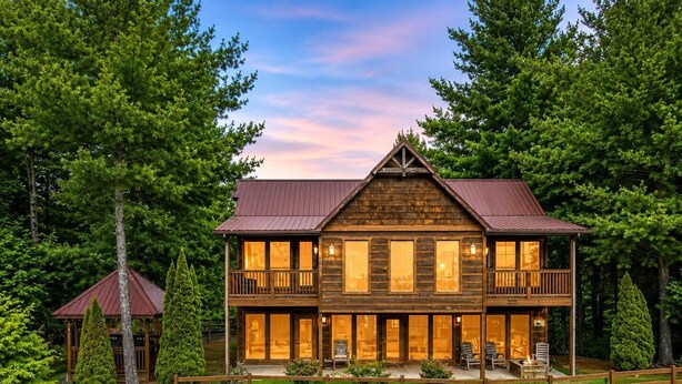 A two-story log cabin with a balcony, surrounded by a wooden fence and lush greenery.