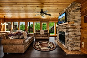A log cabin interior with a stone fireplace, two leather sofas, a ceiling fan, and large windows.