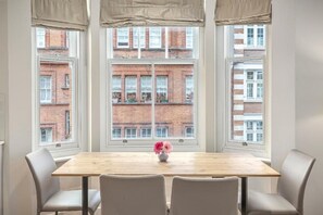 A dining area with a wooden table and four chairs, large windows with a view of brick buildings, and a vase with flowers on the table.