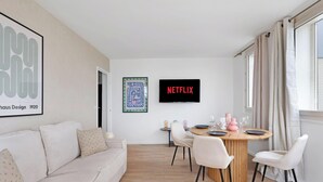 Un salon moderne comprenant un canapé, une table à manger avec des chaises, un téléviseur fixé au mur et une fenêtre avec des rideaux.