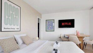 A hotel room with a bed, pillows, a TV, a framed picture, and a table with chairs.