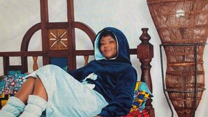 A person lying on a bed with a colorful blanket, a wooden headboard, and a wicker chair in the background. - Kiota Jungle Homestay (Dar es Salaam)