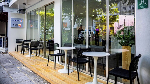 Un espace extérieur aménagé avec des tables et des chaises, une enseigne indiquant « Café Terminal 27 » et un panneau vert portant la mention « Point de rassemblement ».