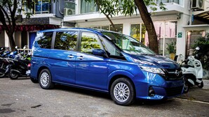 Un monospace bleu est garé dans une rue bordée d’arbres, avec des bâtiments en arrière-plan.