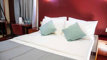 Cama bem arrumada com lençóis brancos e duas almofadas verdes, mesa de cabeceira de madeira com abajur, e parede vermelha com luminária.