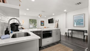 A modern kitchen with a stainless steel sink, built-in oven, and white cabinetry. There is a dining area with a table and chairs, and a large framed picture on the wall. - NEW Soho Sanctuary - Gorgeous Apartment Steps to S Howard Ave With Fenced Yard (Tampa)