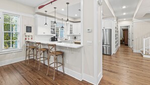 Une cuisine moderne avec des meubles blancs, des tabourets de bar en bois, des luminaires suspendus et un réfrigérateur en acier inoxydable.