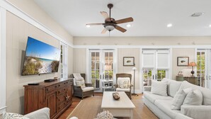 Un salon spacieux doté d’un ventilateur de plafond, d’un grand téléviseur à écran plat, d’un canapé blanc, d’une table basse et d’une cheminée.