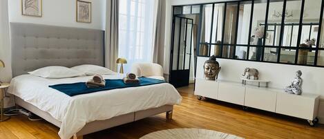A modern bedroom with a large bed, a bedside table, a white console, a round rug, and a large window with curtains.