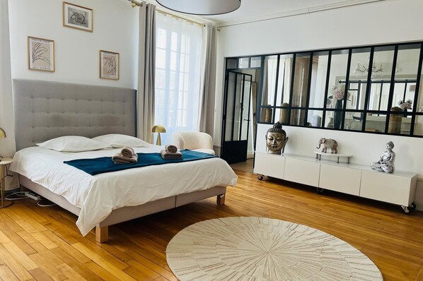 Ein modernes Schlafzimmer mit einem großen Bett, einem Nachttisch, einer weißen Ablage, einem runden Teppich und einem großen Fenster mit Vorhängen.