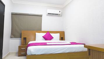 A neatly made bed with a wooden headboard, a bedside table, a window with a blind, and a wall-mounted air conditioning unit.