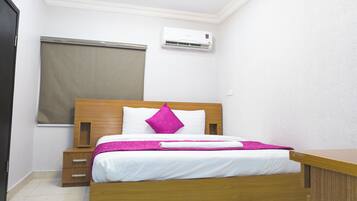 A neatly made bed with a wooden headboard, a white pillow, and a pink accent pillow. There is a wooden nightstand with drawers, a window with a blind, and a wall-mounted air conditioner.