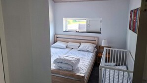 A bedroom with a wooden bed, a crib, a window with a view, and a wall-mounted shelf. - 4 Star Holiday Home in Karrebaeksminde (Karrebæksminde)