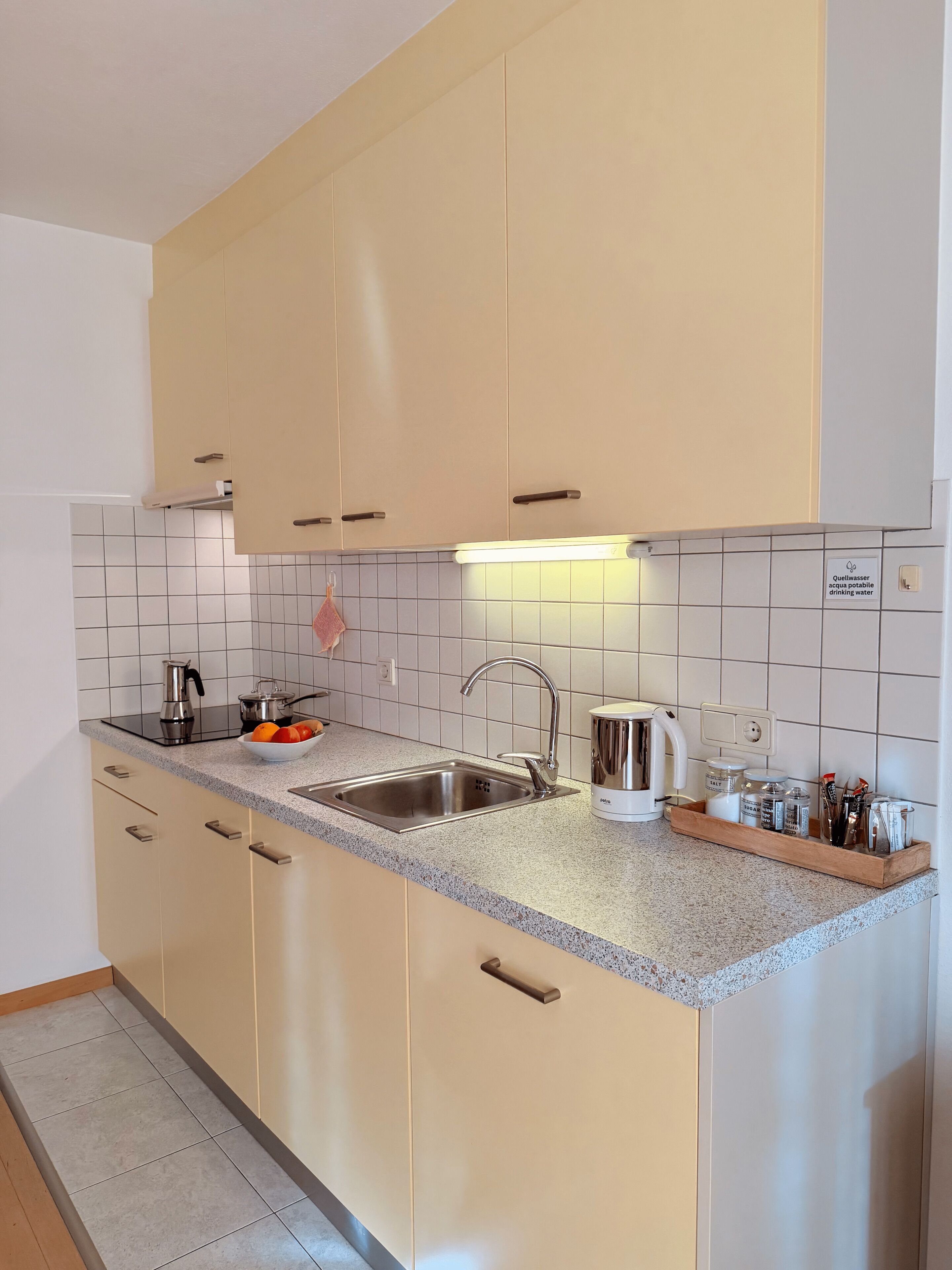 A modern kitchen with beige cabinets, a granite countertop, stainless steel sink, and a kettle.