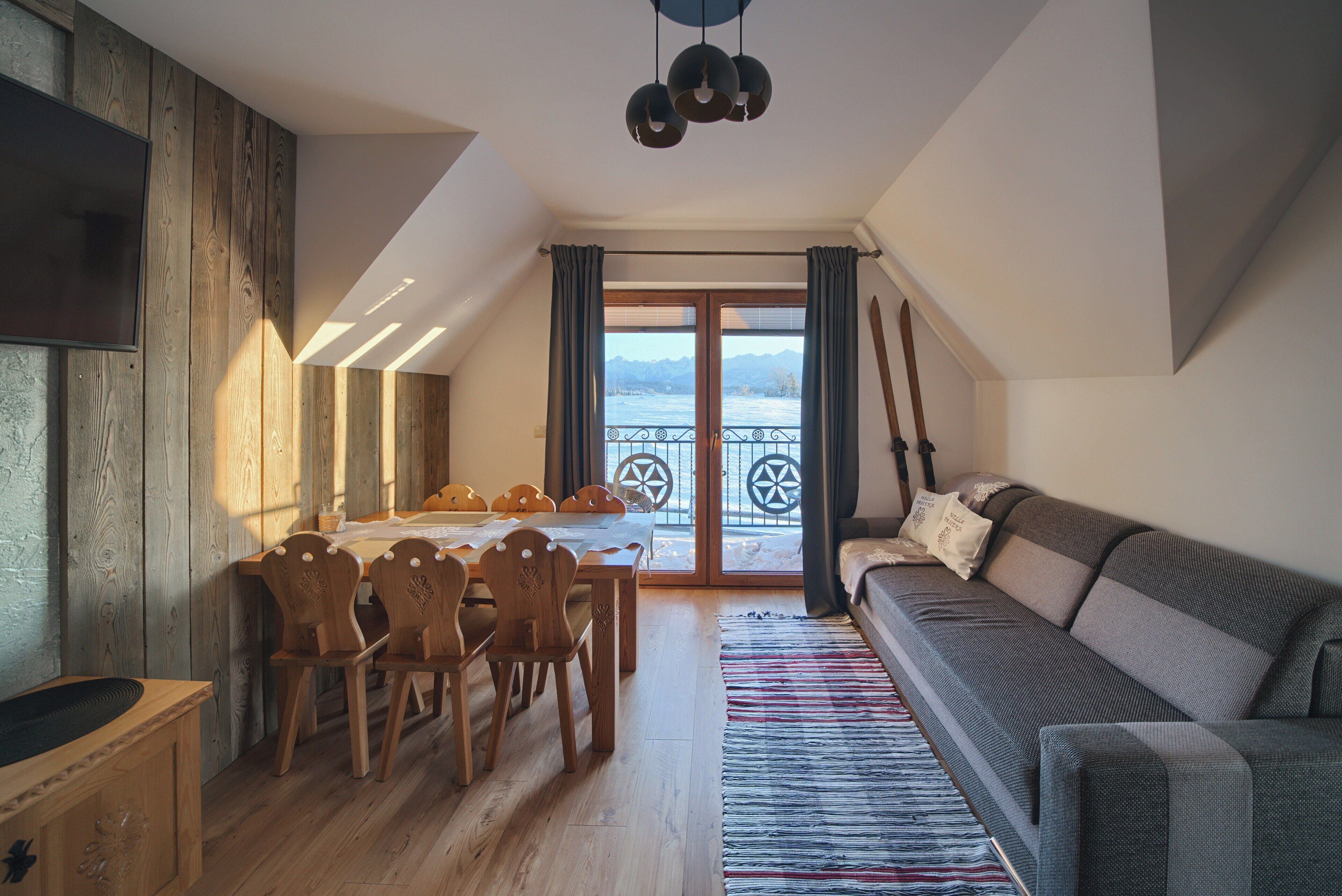 A cozy living room with a sofa, dining table, and chairs. A TV is mounted on the wall, and there's a view of a balcony with a railing.