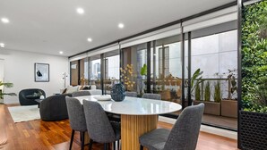 A modern dining area with a round table, grey upholstered chairs, and a wooden base. - 3 Bedroom, Stylish, Spacious Stunner! Huge Balcony, Minutes to Marvel! (Melbourne)
