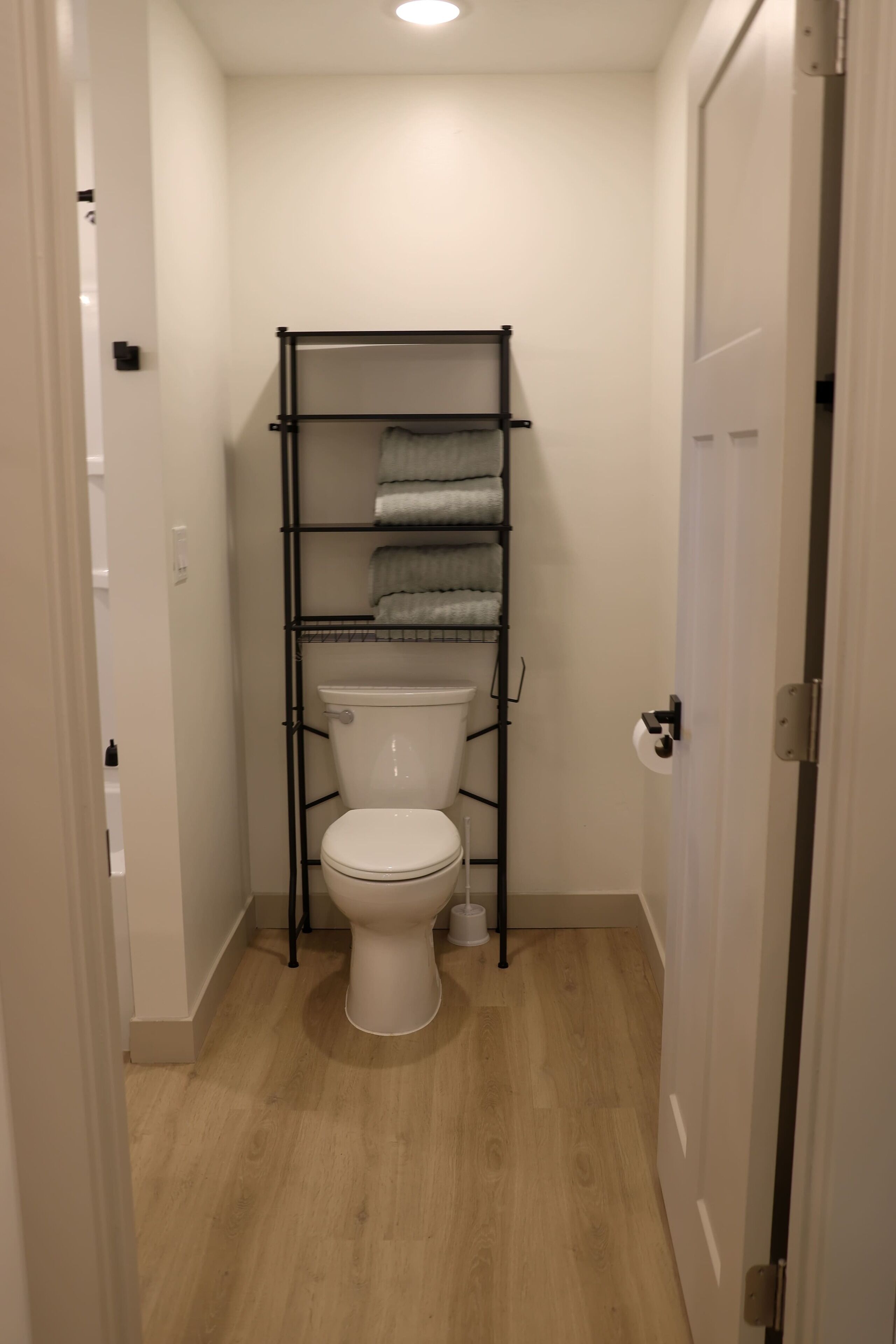 Een kleine badkamer met een wit toilet, een metalen plankenrek met handdoeken en een houten vloer.