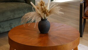 A round wooden coffee table with a dark vase containing dried pampas grass, placed on a patterned rug.