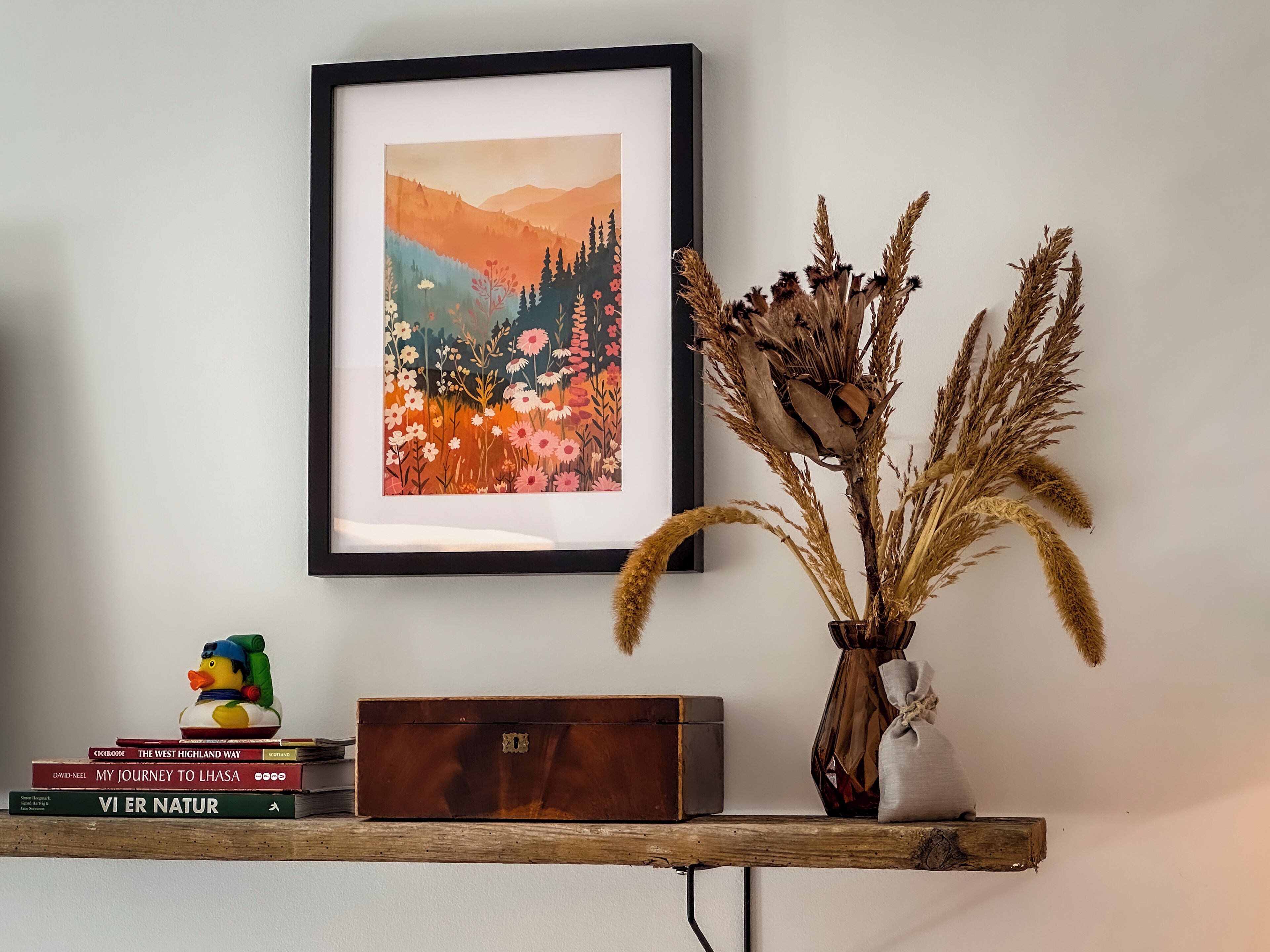 Une œuvre d’art encadrée, une boîte en bois, une figurine de canard et un vase contenant des plantes séchées, le tout posé sur une étagère.
