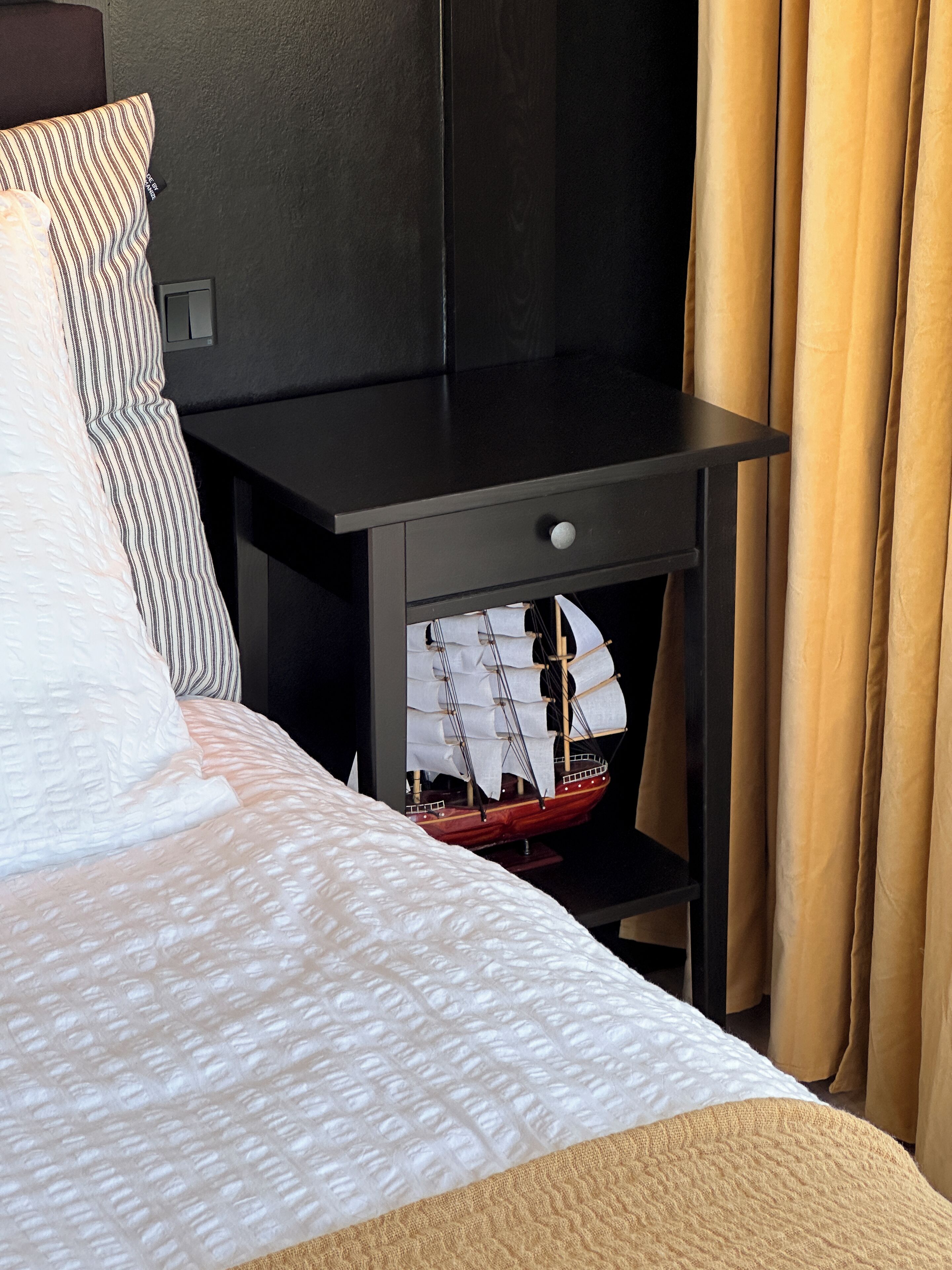 A neatly made bed with patterned bedding, a small black nightstand with a drawer, and a model ship on the nightstand.