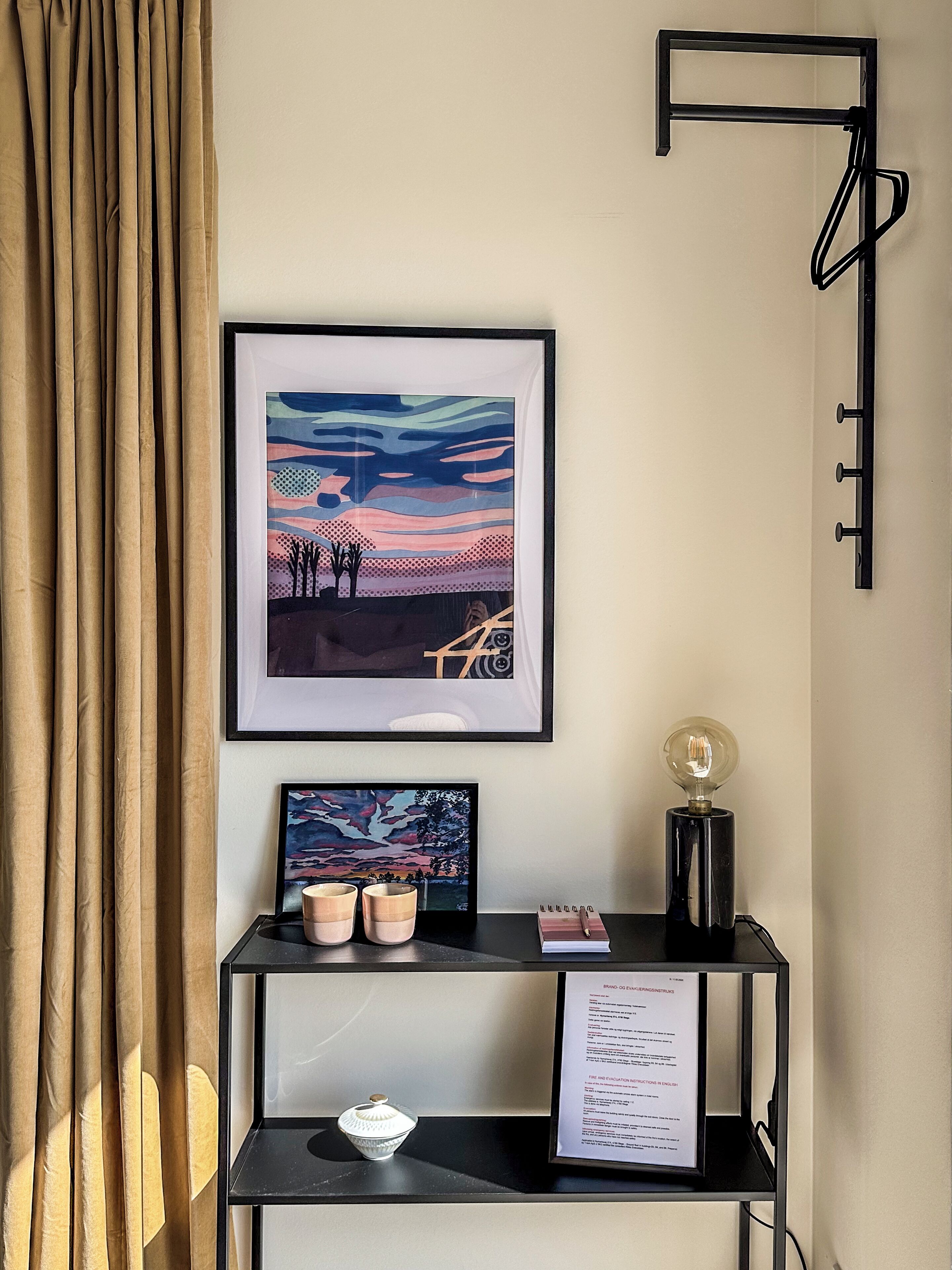 A framed abstract painting, a small desk with a lamp, and a wall-mounted rack.