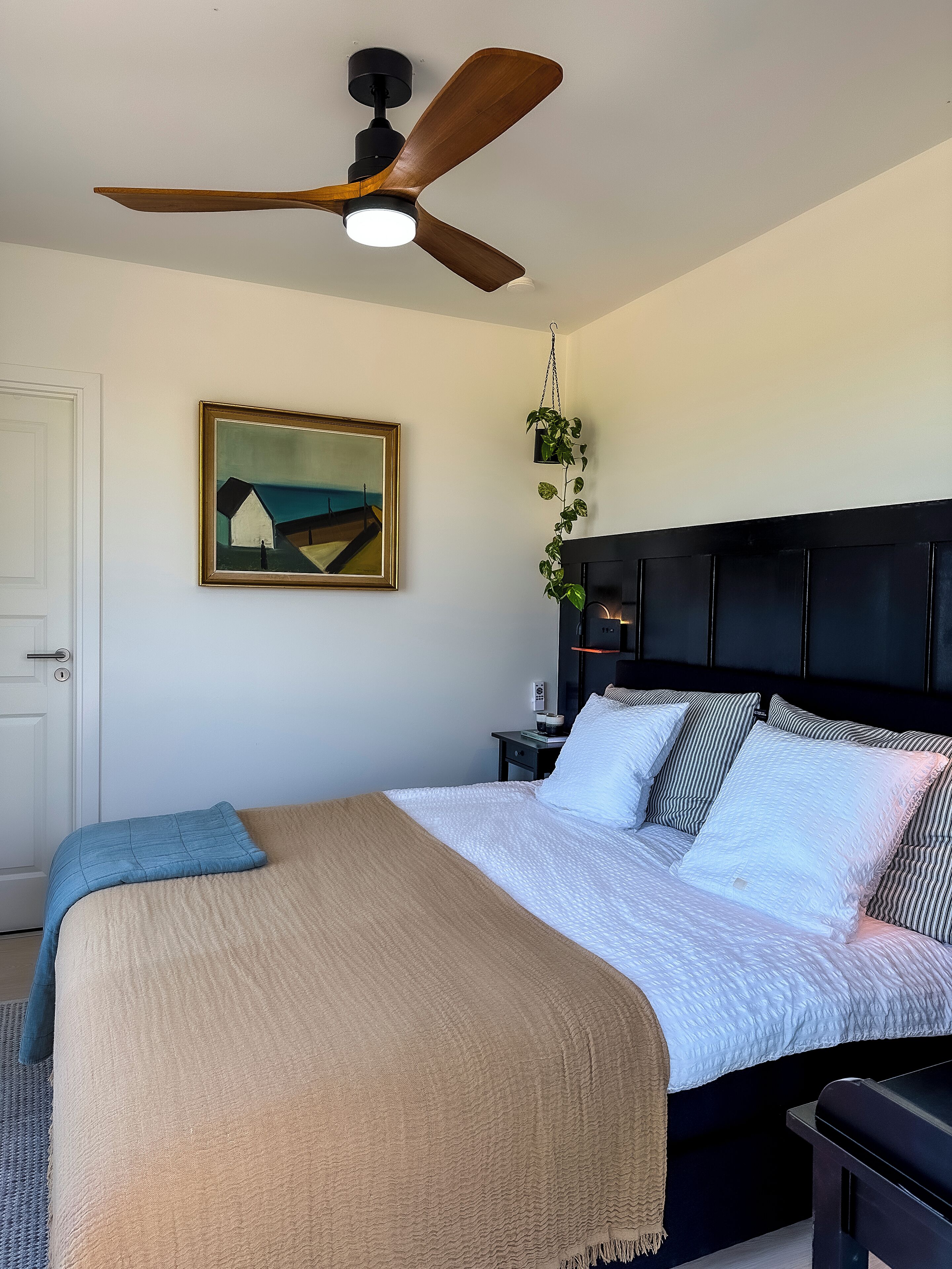 Une chambre à coucher comprenant un lit, un ventilateur de plafond, un tableau, une table de chevet et une commode.