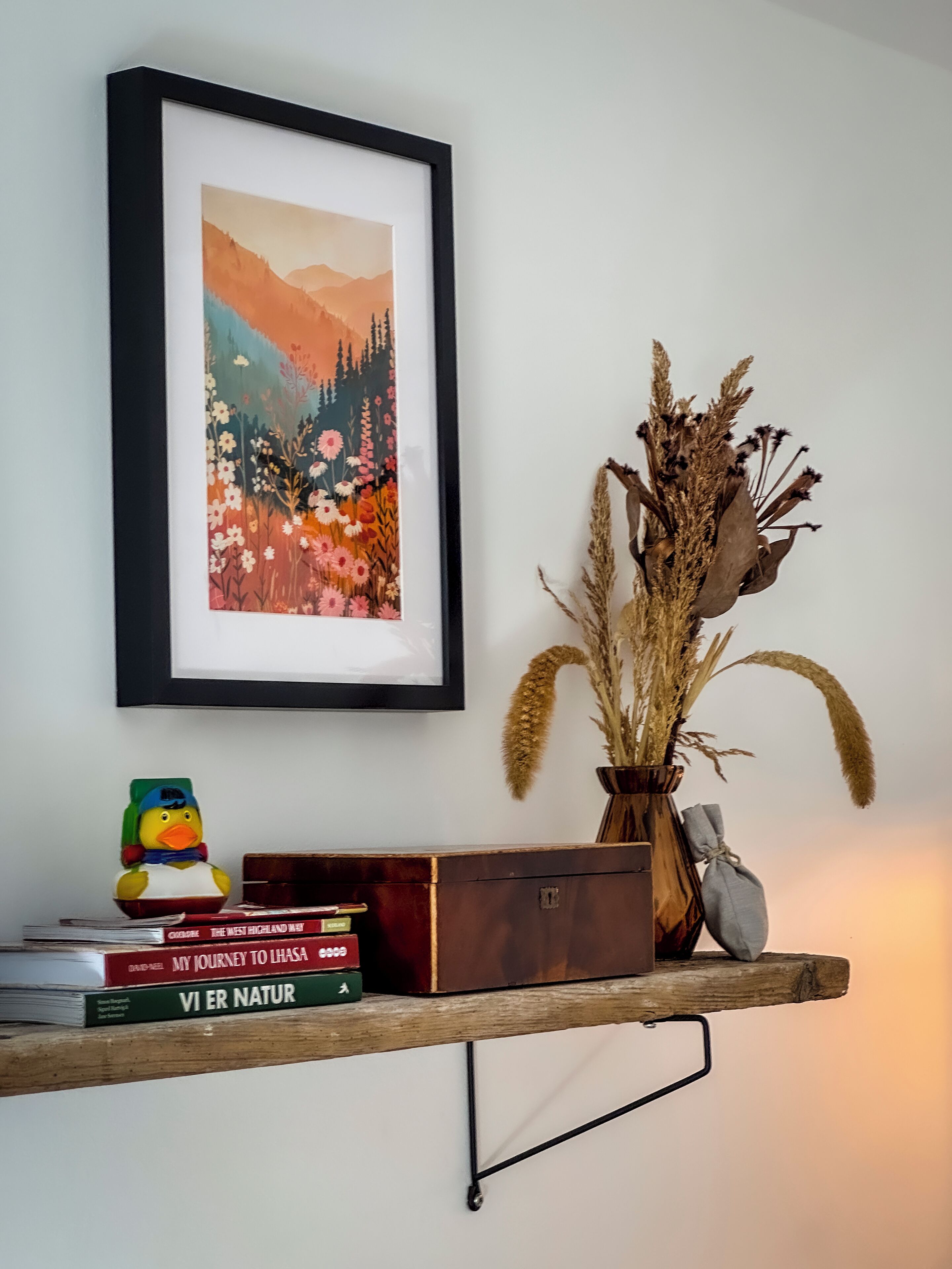 Un cuadro de paisaje enmarco, un jarrón decorativo con plantas secas, un patito de goma y una pila de libros sobre un estante de madera frente a una pared blanca.
