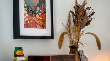 Un cuadro de paisaje enmarco, un jarrón decorativo con plantas secas, un patito de goma y una pila de libros sobre un estante de madera frente a una pared blanca.