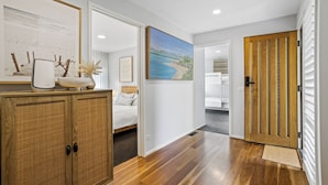 A hallway with wooden flooring, a bed with white bedding, a painting of a beach, and a cabinet with a vase.