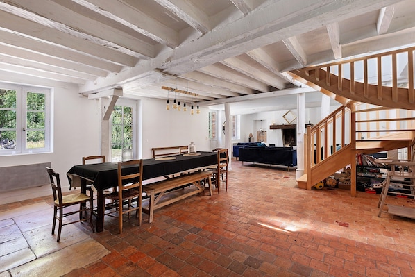 A spacious room with a wooden dining table, chairs, a bench, a staircase, and a brick floor.