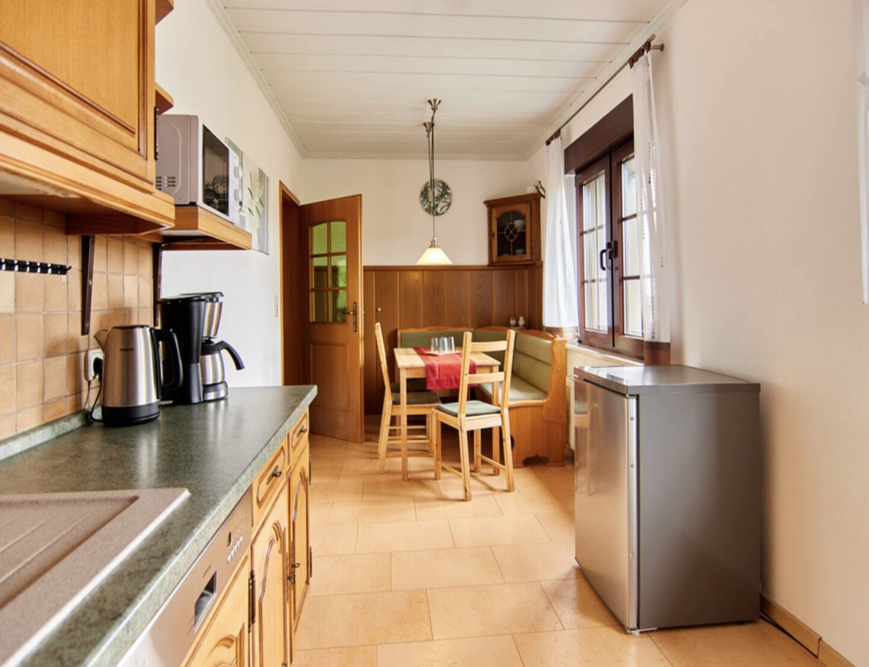 A compact kitchen with wooden cabinets, a dining table with chairs, a refrigerator, and a hanging light fixture.