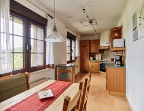 Een keuken met houten kastjes, een eettafel met stoelen, een hanglamp en uitzicht op het groen door grote ramen.