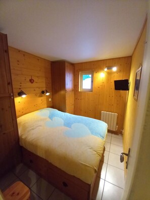 A bedroom with a bed, wooden walls, a small window, a radiator, and a TV mounted on the wall.