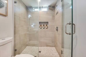 A modern bathroom with a glass-enclosed shower, beige tiles, and a small shelf with bottles.
