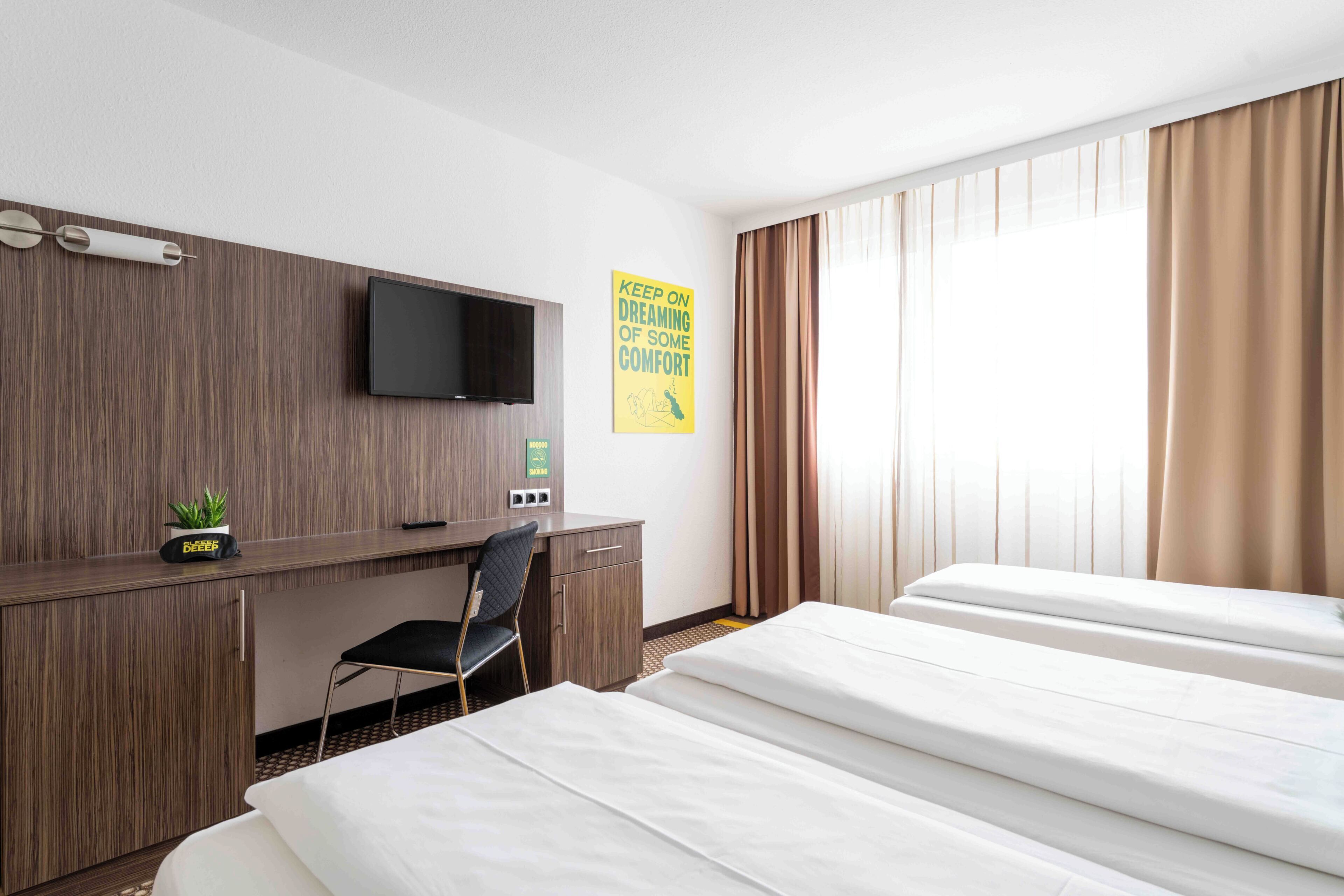 A hotel room with a bed, a desk with a chair, a television, and a poster on the wall.