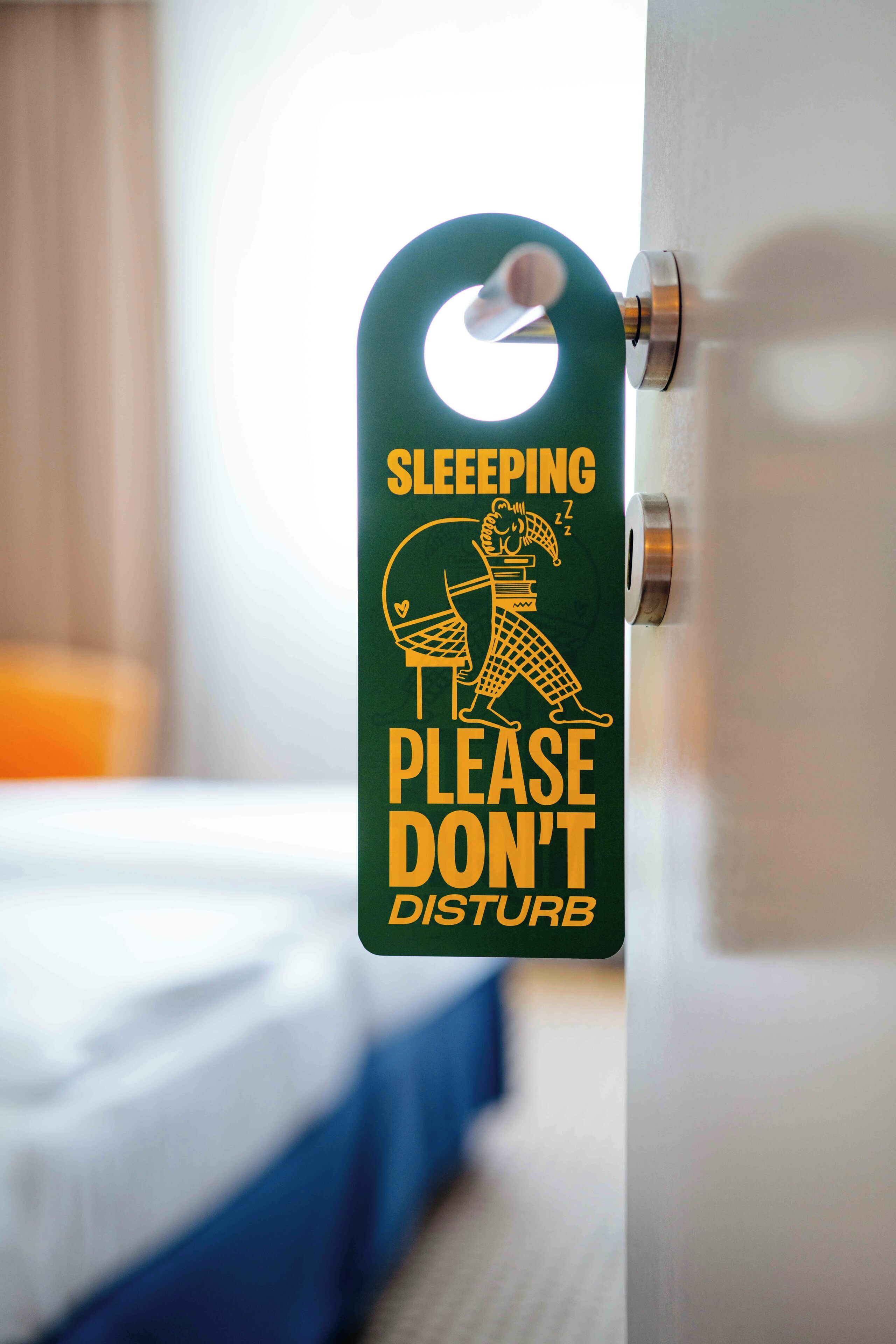 Un letrero verde muestra una persona durmiendo y el texto 'SLEEPING PLEASE DON'T DISTURB' sujeto a un pomo de puerta.