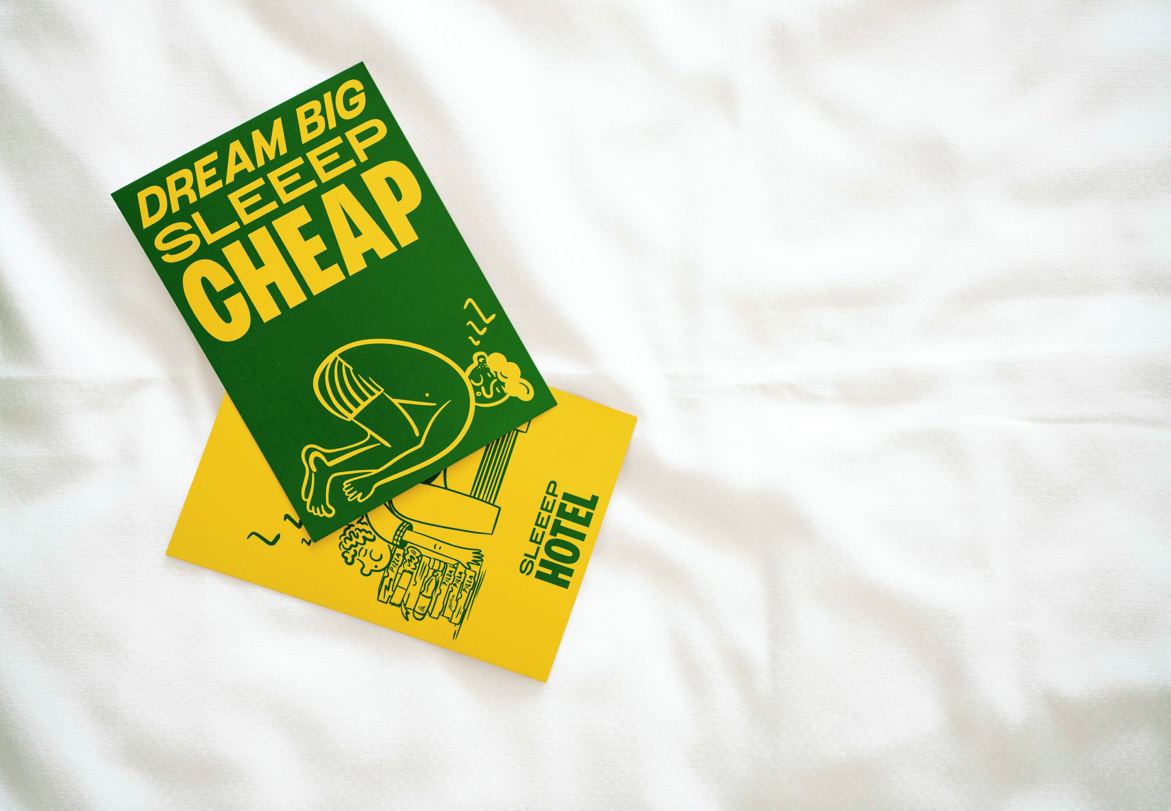 Un libro con el título 'Dream Big Sleep Cheap', que muestra una figura durmiendo e una ilustración de un hotel sobre un fondo blanco.