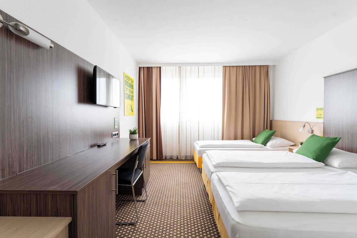 A hotel room with a desk, two beds, a TV, and a large window with curtains.
