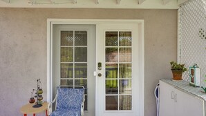 A covered porch with a white door, a blue chair, a small table with decorative items, and a cabinet with a plant on top. - Cozy Couple's Oasis w/ Pool: 3 Mi to Disneyland! (Garden Grove)