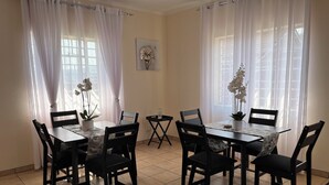 A dining area with a table set for four, surrounded by chairs, with a window and curtains on one side and a small table with a plant on the other. - Soothing 4-bedroom house in Manzini (Manzini)
