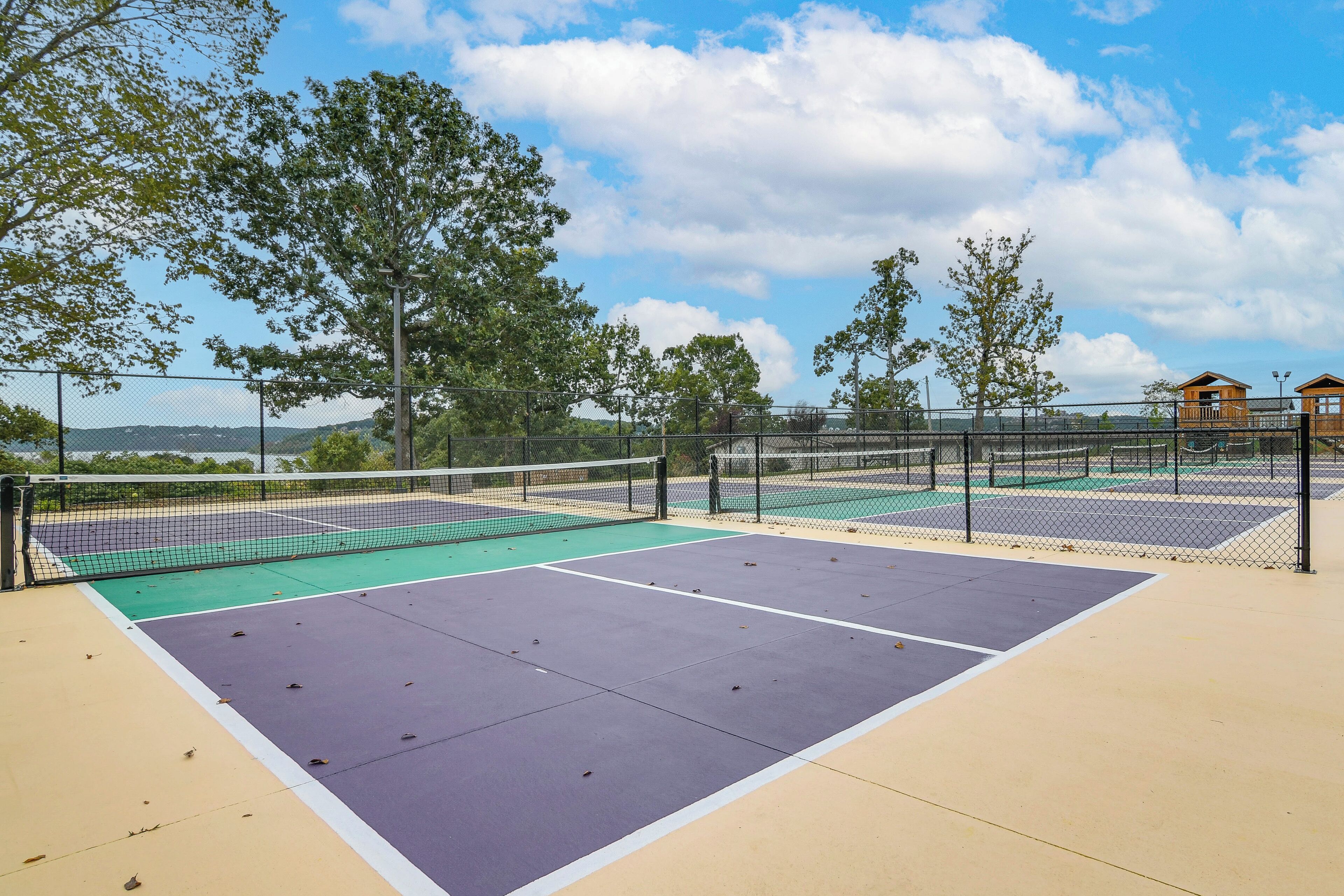 An outdoor tennis court with a net, surrounded by a fence and trees.