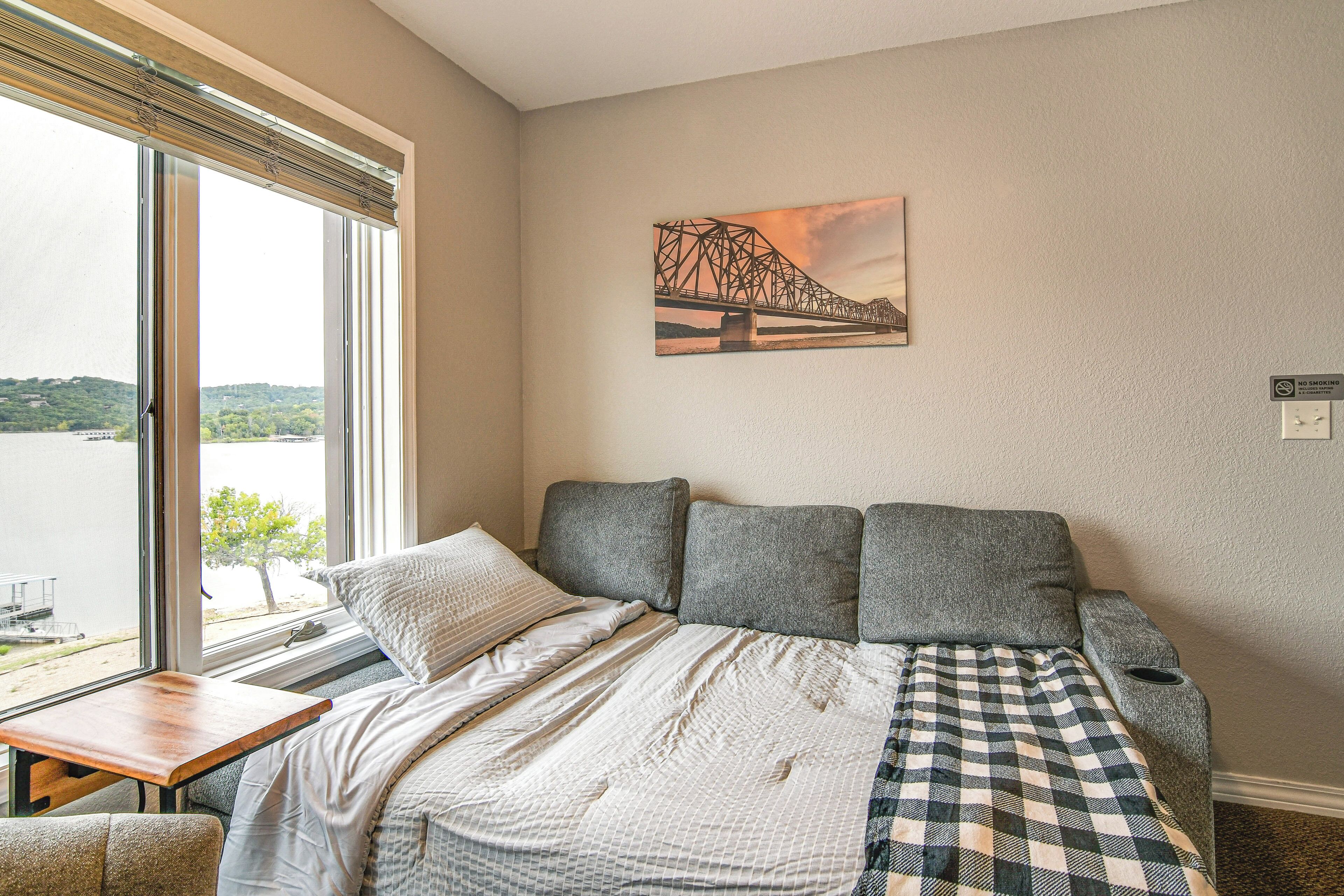 A bedroom with a large window, a bed with a checkered blanket, and a painting of a bridge on the wall.