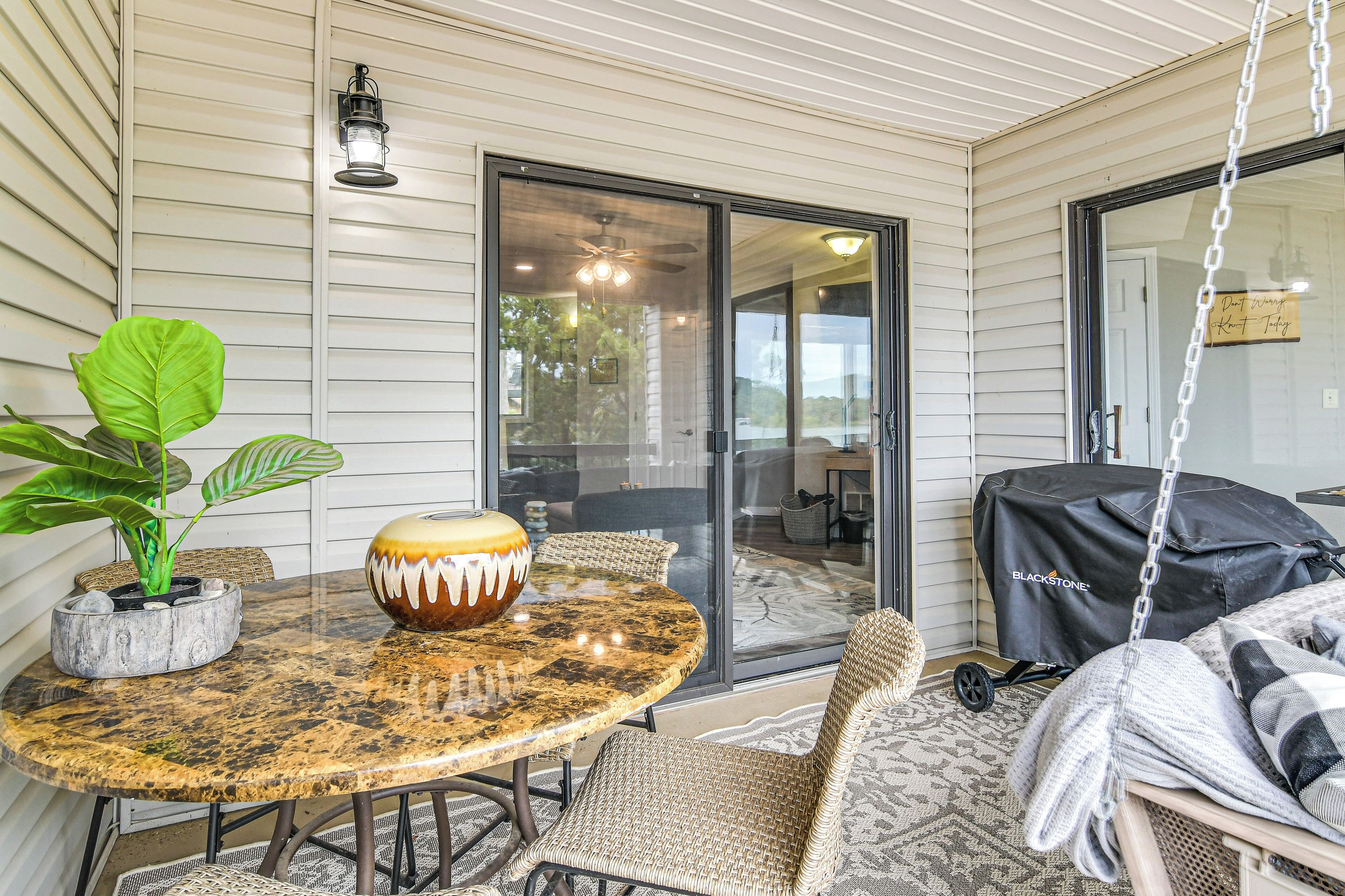 A covered patio area with a round table, wicker chairs, a hanging chair, a potted plant, and a grill.