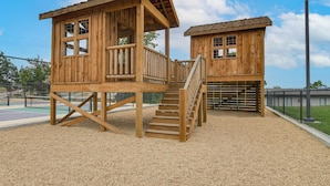 Two wooden playhouses with stairs and railings, surrounded by a playground area with a fence and a sports court. - Table Rock Lake Retreat: Steps to Water & Pools! (Kimberling City)