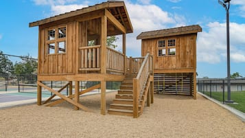 Dos casitas de juegos de madera con escaleras y barandas, ubicadas en un área de juegos con valla y cancha deportiva.