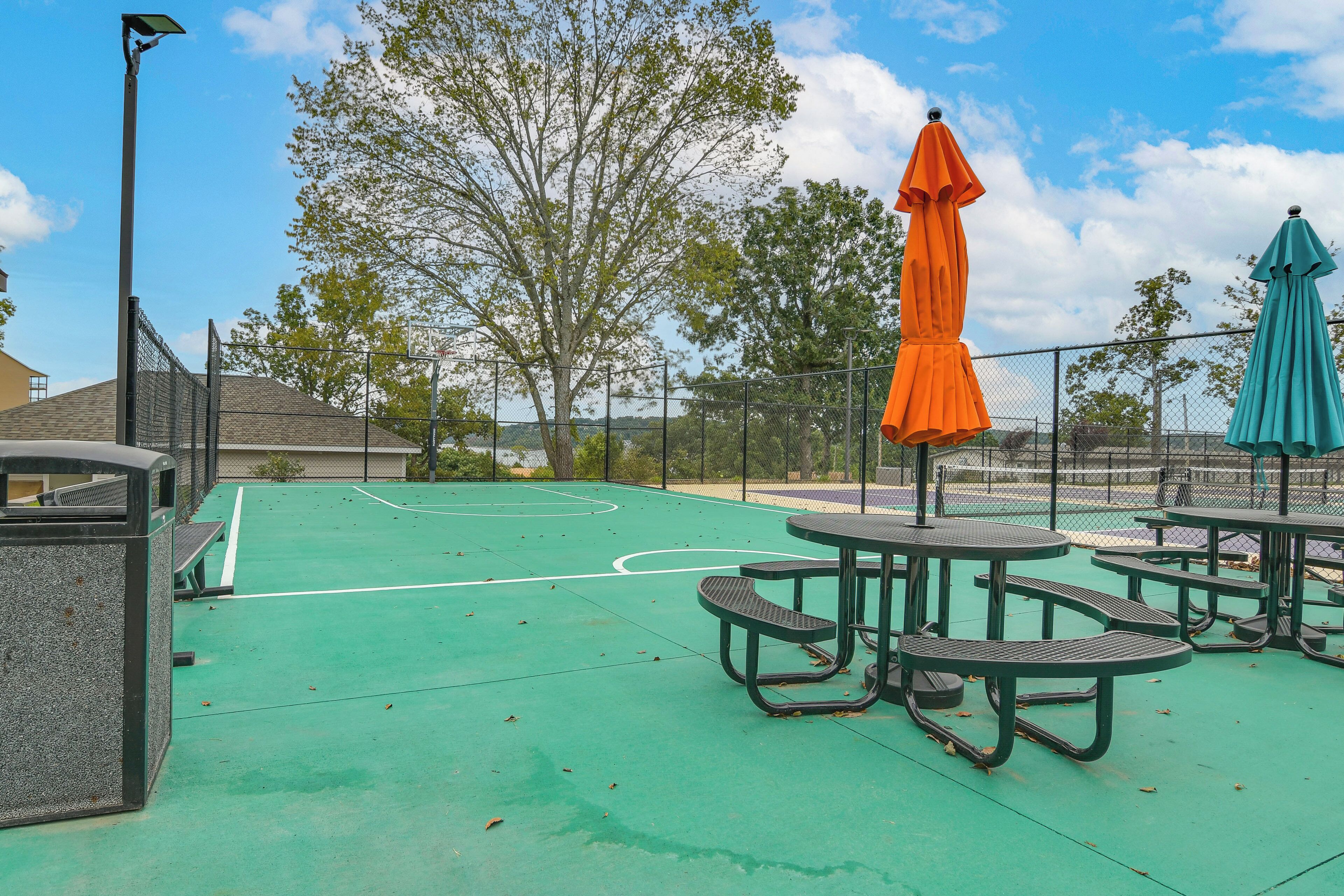 Un terrain de basket en plein air, équipé de tables de pique-nique et de parasols, entouré d’une clôture et d’arbres.